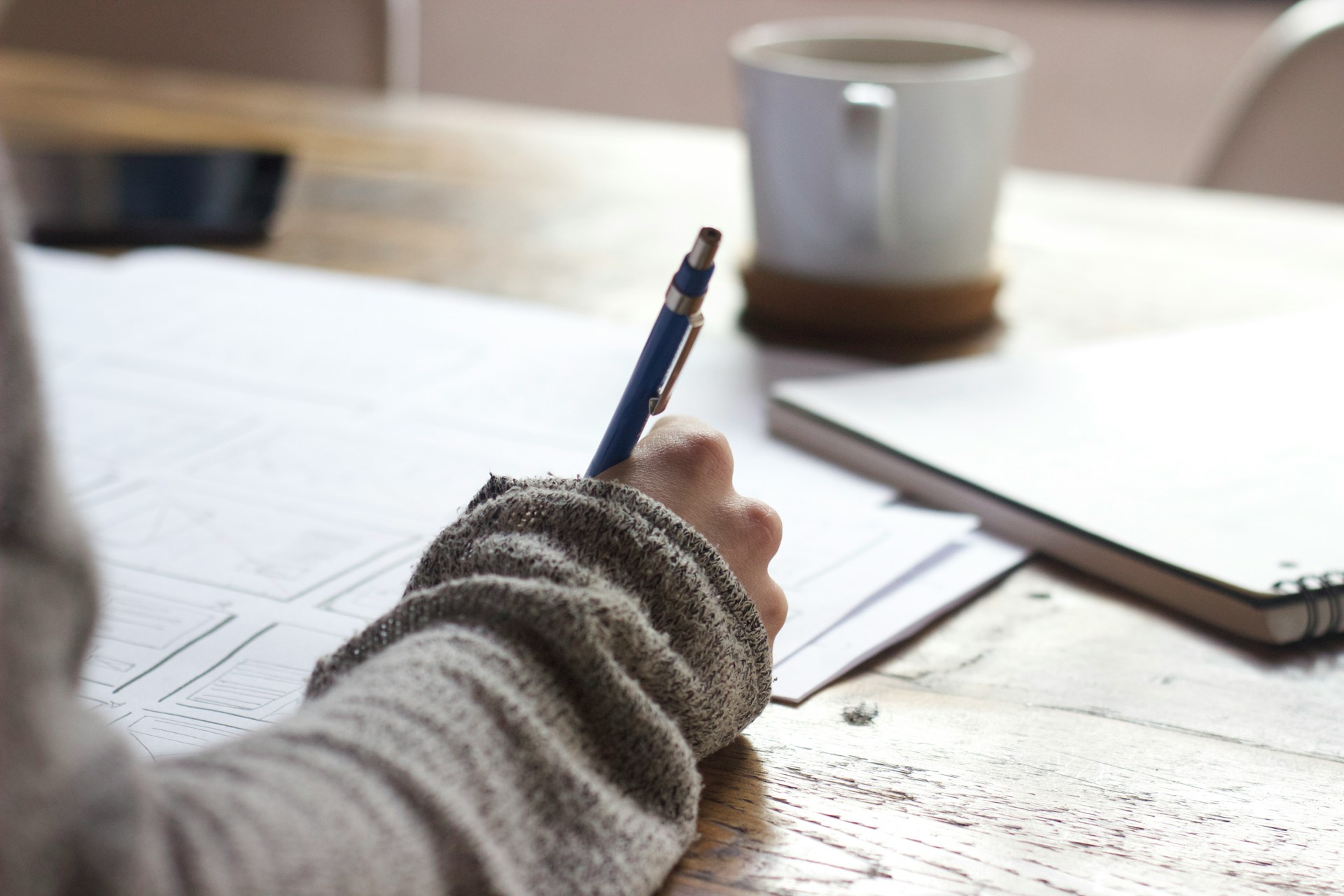 person writing on papers with a ceramic coffee mug on wooden table