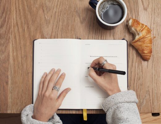 woman writing in a notebook planner with a coffee and croissant on the table