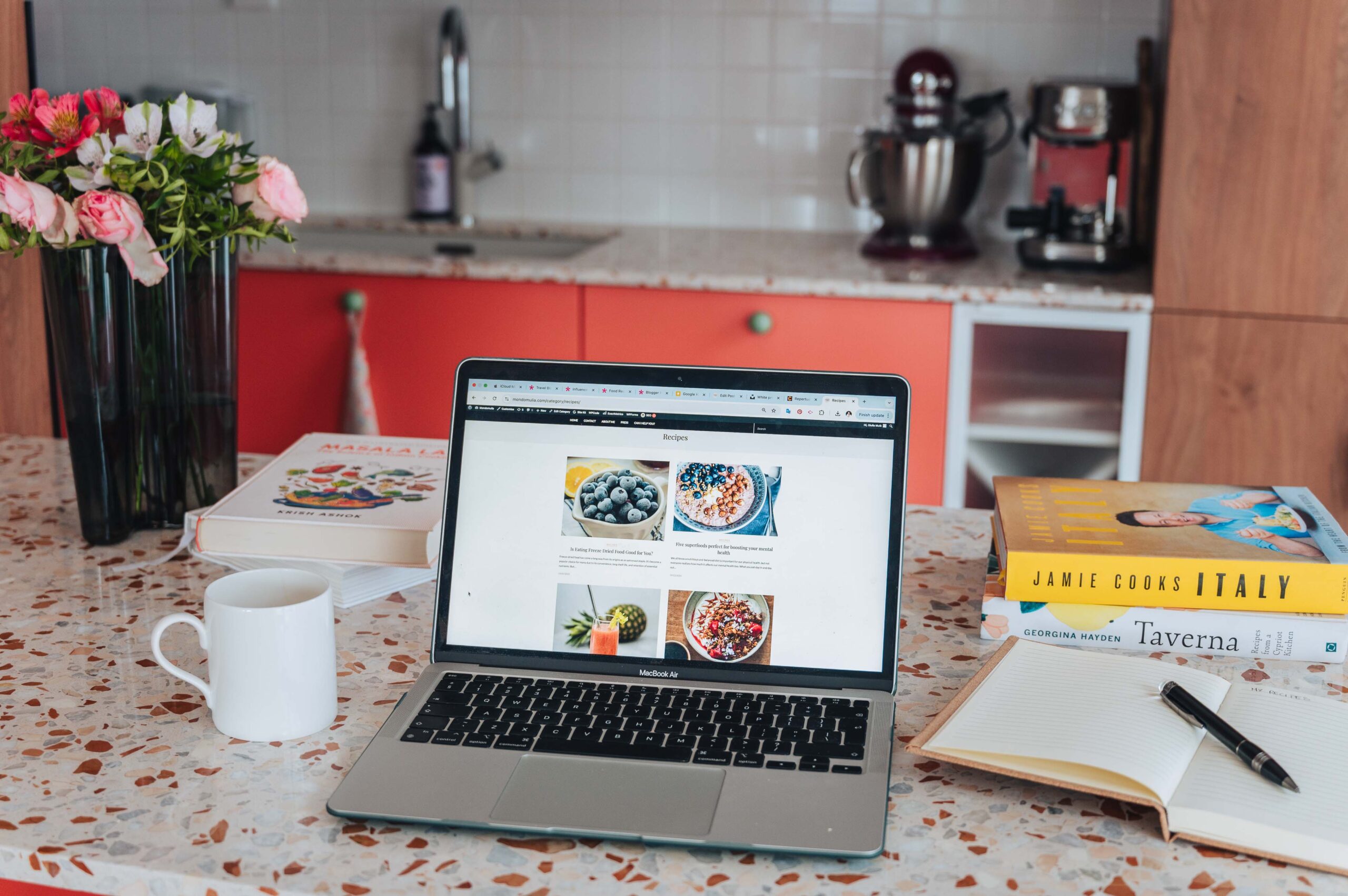 Laptop on a marble kitchen counter with a coffee cup, flowers and cookbooks