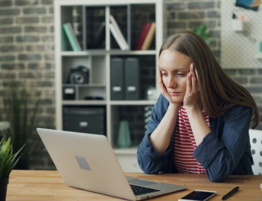 Tired young woman using laptop in office touching head feeling headache