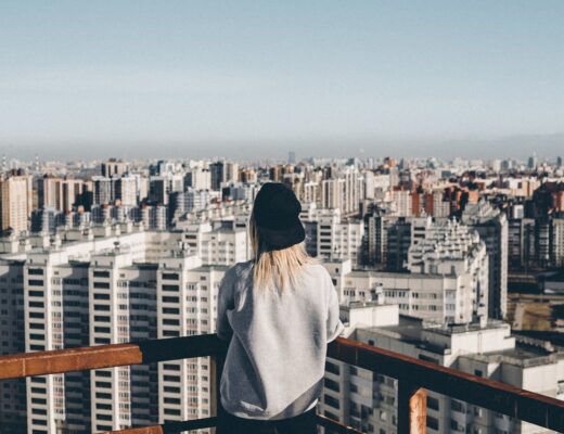 woman in grey sweatshirt and black hat standing on top of building overlooking big city