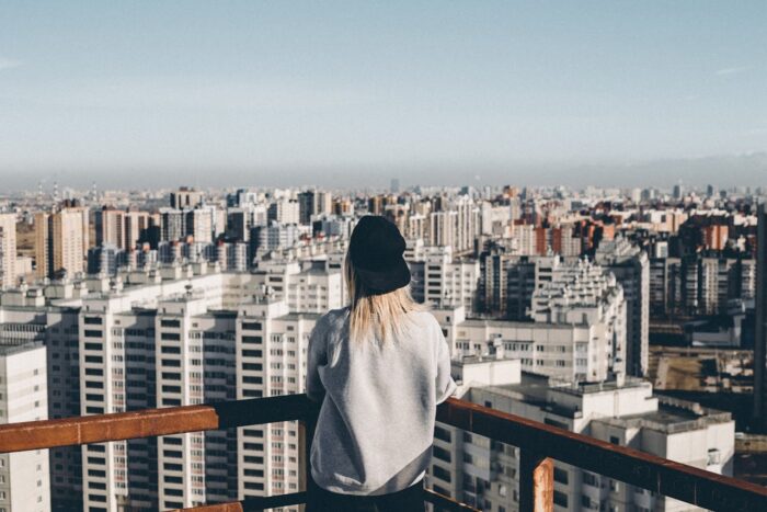 woman in grey sweatshirt and black hat standing on top of building overlooking big city