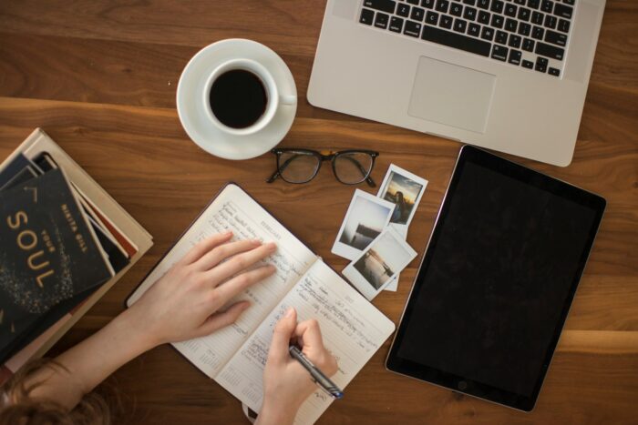 person writing a travel journal holding a pen with a coffee and computer on the desk person writing a travel journal holding a pen with a coffee and computer on the desk