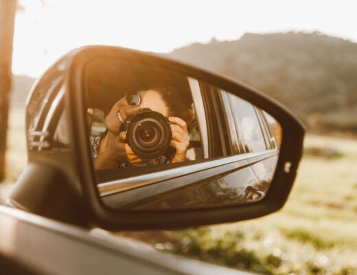 A young woman takes a photo through a car mirror reflection, capturing a sunny outdoor scene.
