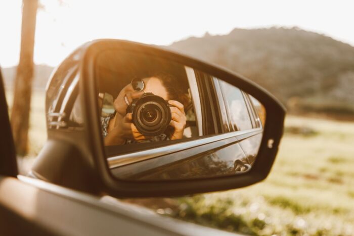 A young woman takes a photo through a car mirror reflection, capturing a sunny outdoor scene. A young woman takes a photo through a car mirror reflection, capturing a sunny outdoor scene.