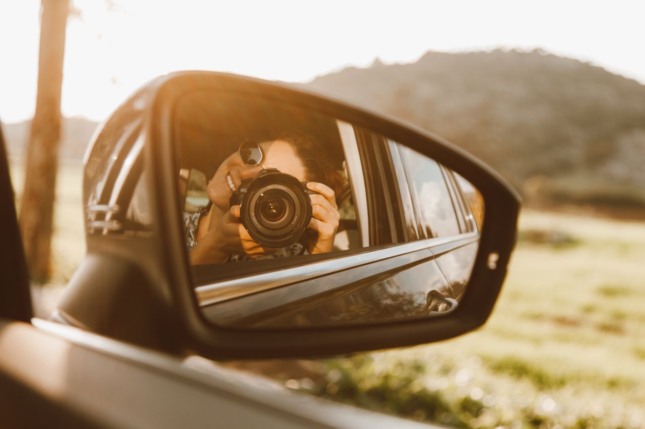 A young woman takes a photo through a car mirror reflection, capturing a sunny outdoor scene.