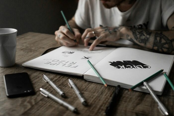 Man with tattoos and white shirt sketching with a black pen on a white notebook Man with tattoos and white shirt sketching with a black pen on a white notebook